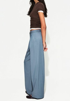 Nainen seisoo sivuttain, yllään ruskea lyhythihainen paita ja leveälahkeiset siniset raidoitetut housut, jalassa tummat kengät, valkoista taustaa vasten.