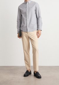 Muž v svetle šedé košili na knoflíky, béžových kalhotách a černých mokasínech, držící černou peněženku, stojící na šedé podlaze proti bílé zdi.