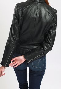 Černá kožená motocyklová bunda s přiléhavým střihem, zipy na manžetách a detaily na ramenou. Nošena přes tmavé džínové kalhoty s prošitím na zadních kapsách.