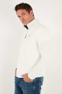 Kreem värvi lukuga fliispullover kõrge kraega, kontrastse sisekraega ja väikese logoga rinnal. Kombineeritud siniste teksadega.