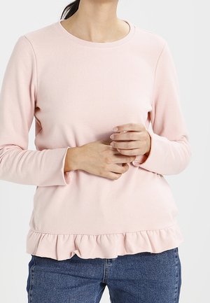 Lichtroze lange mouwen top met een ribbelkraag en een ruffles onderaan. De stof lijkt zacht en gestructureerd, gedragen over donkerblauwe spijkerbroeken.
