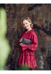 Červené, volánkové šaty s dlouhými rukávy, vrstvový design; černá kabelka s nenápadným logem. Pozadí tvoří texturovaný, tmavý povrch s přírodními odstíny.