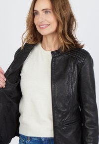 Svart läderjacka med en slät textur, med dragkedjestängning och quiltade axeldetaljer, bärs över en ljus tröja och blå jeans.