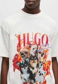 T-shirt bianco in cotone con una grafica colorata che mostra diversi cani e la parola "HUGO" in rosso bold, circondata da fiamme arancioni.