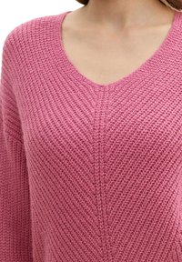 Maglione rosa lavorato a maglia con design a V. Presenta un motivo testurizzato e una vestibilità ampia, che mette in evidenza un tessuto morbido e caldo.