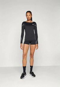 Mujer de pie vistiendo una camiseta deportiva negra de manga larga, pantalones cortos negros, calcetas negras y zapatillas Adidas negras contra un fondo claro y simple.