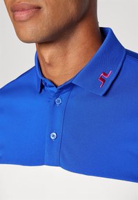 Blå pikétröja med en strukturerad krage, två blå knappar och en röd logotyputstickning på kragen. Slät, lätt tyg.