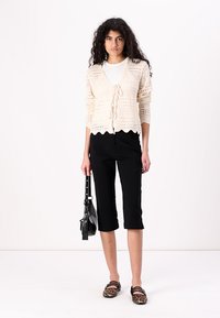 Krämfärgad virkad kofta med vågig fåll, knuten i midjan; matchad med svarta kapri-byxor och leopardmönstrade ballerinas, håller en svart handväska.