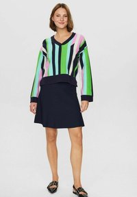 Barvna pristrižena pulover v zeleno, rožnato, modro in črno, v kombinaciji z mornarsko modro krilom v stilu skater. Mehka tkanina, V-izrez, dolgi rokavi.