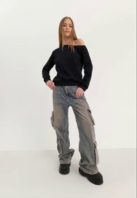 Sudadera negra de hombros descubiertos combinada con pantalones cargo holgados de color azul claro que tienen bolsillos laterales y calzado oscuro y grueso. Fondo simple.