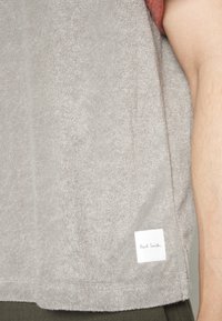 Vaaleanharmaa froteepaita, jossa on valkoinen etiketti, jossa lukee "Paul Smith" helman kohdalla, näkyvää ompelua ja rento istuvuus.