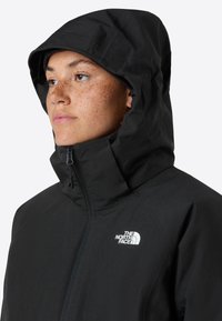 Giacca nera impermeabile con colletto alto e cappuccio attaccato. Presenta una zip frontale e un piccolo logo bianco sul petto. Texture liscia.