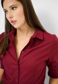 Camicia bordeaux con maniche corte, dotata di un colletto classico, texture liscia e due bottoni frontali.