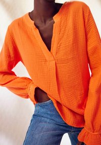 Camicetta arancione a maniche lunghe in tessuto testurizzato con scollo a V e polsini arricciati, abbinata a jeans in denim blu slim.