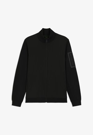 Svart jakke med høy krage, glidelåslukking, sidelommer, ribbestrikkede mansjetter og en glidelåslomme på venstre erme. Jevn stofftekstur.