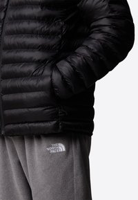 Personne portant une veste noire matelassée, les mains dans les poches, et un pantalon en polaire gris avec le logo The North Face sur la cuisse.