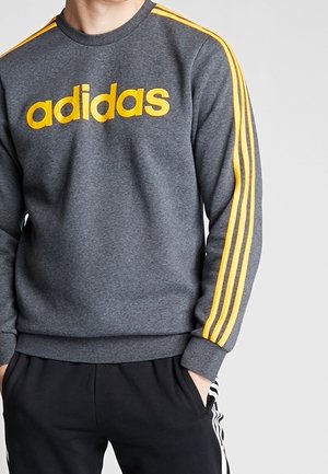 Osoba nosząca szarą bluzę Adidas z żółtymi paskami na rękawach i żółtym logo, w połączeniu z czarnymi spodniami Adidas z białymi paskami.