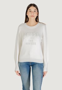 Crème sweater met ribbels aan de onderkant en mouwen,Featuring een zilveren "GUESS" logo en oprichtingsdatum, gedragen met blauwe jeans.