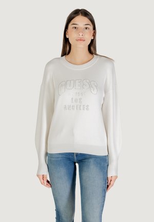 Crème sweater met ribbels aan de onderkant en mouwen,Featuring een zilveren "GUESS" logo en oprichtingsdatum, gedragen met blauwe jeans.