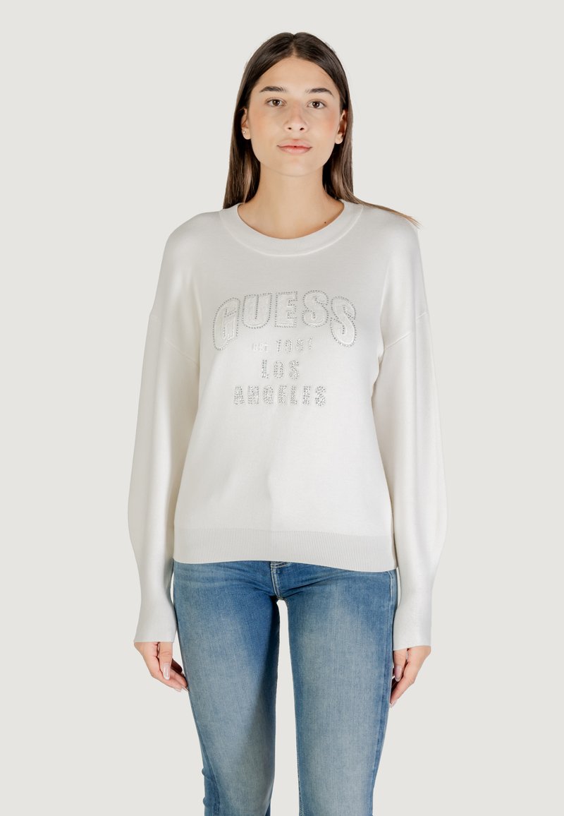 Pull crème avec ourlet et poignets côtelés, affichant un logo "GUESS" argenté et une date de création, porté avec un jean bleu.