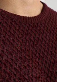 Burgundy stickad tröja med ett texturerat diamantmönster, ribbad halslinning och mjukt material; närbild av tyget.