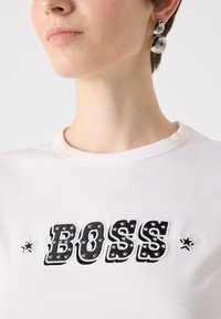 Balta medvilninė marškinė su užrašu "BOSS" juoda spalva ir žvaigždžių raštais, apvaliu iškirpimu ir trumpomis rankovėmis. Išvaizdą papildrina sidabrinės auskarų smeigtukai.