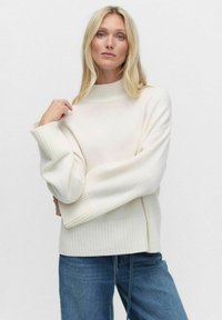 Krämfärgad tröja med hög halsringning, ribbade ärmslut och en avslappnad passform. Mjuk textur, kombinerad med blå denimjeans.