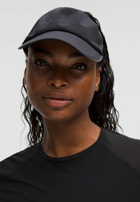 Jonge zwarte vrouw met lang nat haar, die een donkere sportpet en een zwart sportshirt draagt, en zelfverzekerd vooruit kijkt op een effen grijze achtergrond.