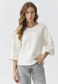 Witte V-hals trui met brede driekwartmouwen, gemaakt van een zachte stof. Paar grijze jeans met een relaxte pasvorm.