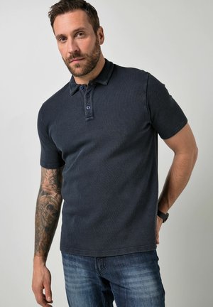 Mann mit tätowiertem Arm, der ein dunkelnavyfarbenes Poloshirt und blaue Jeans trägt, steht mit einer Hand hinter dem Rücken vor einem einfarbigen Hintergrund.