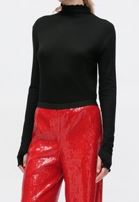 Personne portant un haut noir à col roulé ajusté et un pantalon rouge à paillettes taille haute et brillant, debout devant un fond clair et uni.