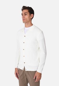 Cardigan bianco a coste con chiusura a bottoni, dotato di bottoni marroni e colletto classico. Indossato sopra una camicia bianca, abbinato a pantaloni marroni.