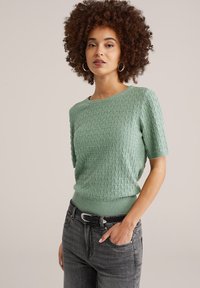 Miętowo zielony sweter z krótkim rękawem, wykonany na drutach, z teksturowanym geometrycznym wzorem i ściągaczem u dołu. Połączony z szarymi jeansami z wysokim stanem.