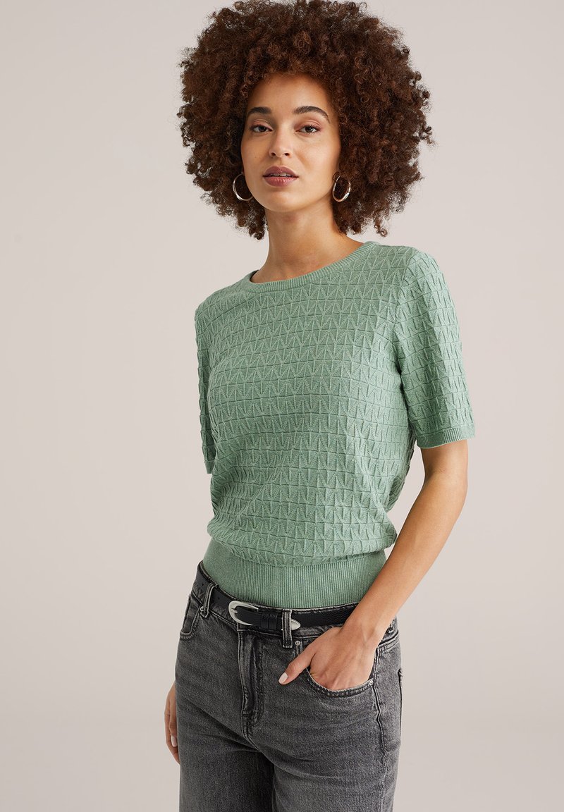 Miętowo zielony sweter z krótkim rękawem, wykonany na drutach, z teksturowanym geometrycznym wzorem i ściągaczem u dołu. Połączony z szarymi jeansami z wysokim stanem.