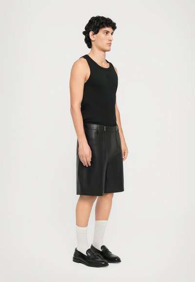 Man standing in black sleeveless top, black knee-length leather shorts, white socks, and black loafers against a plain white background.
