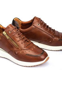 Zapatillas de cuero marrón con cordones, que cuentan con una cremallera lateral, detalles de tachuelas, una suela de goma blanca y un patrón texturizado en la superficie.