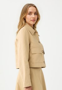 Beige cropped Jacke aus glattem Stoff, mit einem Kragen, Frontknöpfen und zwei großen Brusttaschen. Lange Ärmel und minimale Nähdetails.