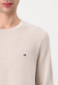 Pull en tricot beige clair avec un col rond. Présente un petit logo tricolore sur la poitrine, mettant en valeur un tissu texturé et respirant.