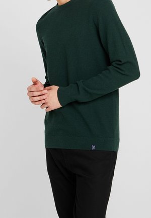 Mann trägt dunkelgrünen, strukturierten Pullover und schwarze Hose, die Hände vor dem Bauch aneinandergereiht vor schlichtem grauem Hintergrund.