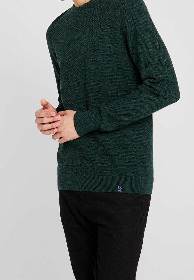 Homme portant un pull vert foncé texturé et un pantalon noir, les mains jointes à la taille sur un fond gris uni.