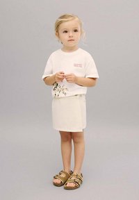 T-shirt blanc à manches courtes avec un imprimé coloré, associé à une jupe en denim crème et des sandales marron avec des accents tressés. Fond gris.