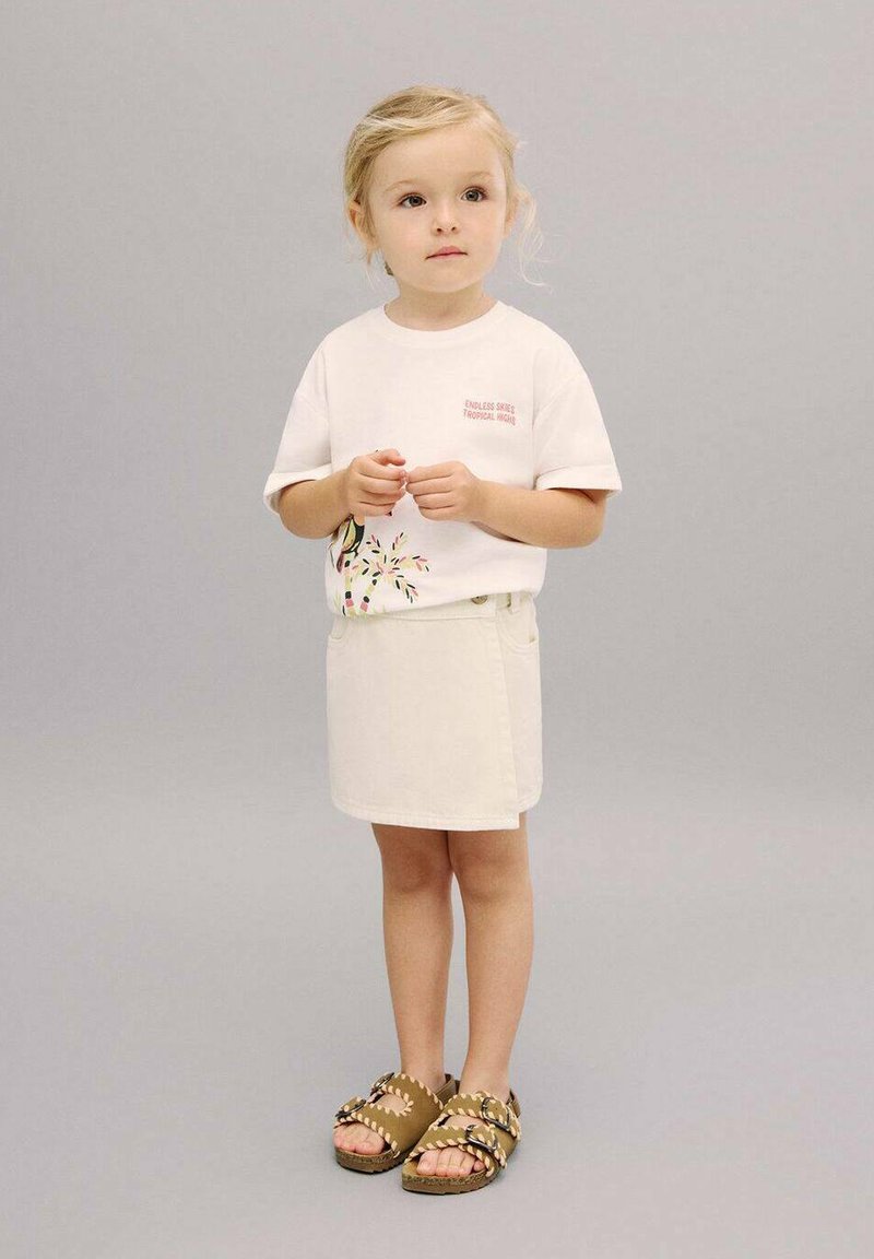 T-shirt blanc à manches courtes avec un imprimé coloré, associé à une jupe en denim crème et des sandales marron avec des accents tressés. Fond gris.