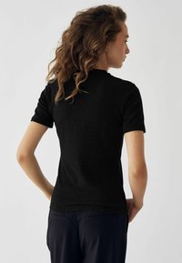 Fekete, rövid ujjú felső, testhezálló fazonnal, texturált anyaggal és kerek nyakkivágással. Háttal látható, vállig érő hajjal és sima szilhuettel.