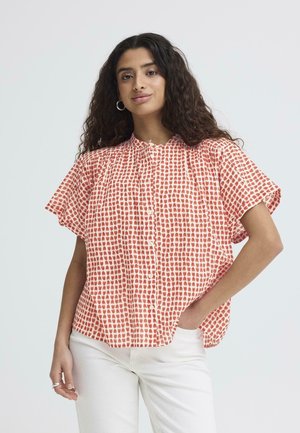 Femme aux cheveux bouclés et foncés portant des boucles d'oreilles en anneaux, un chemisier à manches courtes rouge et blanc à motifs, et un pantalon blanc, debout devant un fond uni.