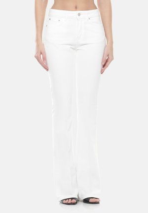 Donna che indossa pantaloni bianchi a vita alta con chiusura a bottone e cerniera, in piedi a piedi nudi su uno sfondo bianco.