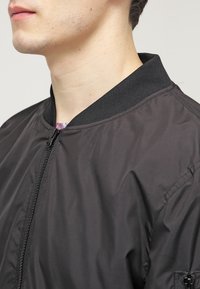 Must have bomber-jakk musta värviga, millel on luku ees, soonikkoes krae ja sile tekstuur. Ilma nähtavate mustrite või metalldetailideta.