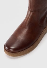 Bota de cuero marrón hasta el tobillo con un acabado suave, punta redondeada y suela de goma texturizada. Presenta una costura sencilla a lo largo del costado.