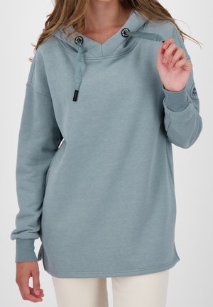 Femme portant un sweat à capuche ample bleu clair avec des cordons, tenant un cordon, associée à un pantalon blanc sur un fond uni.
