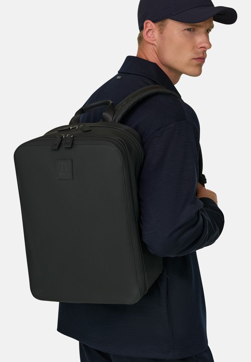 Homme portant une casquette noire et une chemise marine, portant un grand sac à dos noir rectangulaire dans le dos, vu de côté.