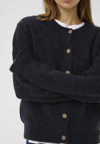 Tmavě šedý, huňatý kardigan s kulatým výstřihem, zapínáním na knoflíky a dlouhými rukávy. Obsahuje světle hnědé knoflíky a texturovaný, měkký materiál.